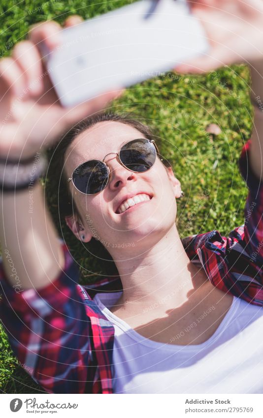 Oben die junge lächelnde Hipsterfrau, die an einem sonnigen Tag im Park auf Gras liegt und dabei mit dem Handy ein Selfie nimmt. obere Ansicht Junge Frau