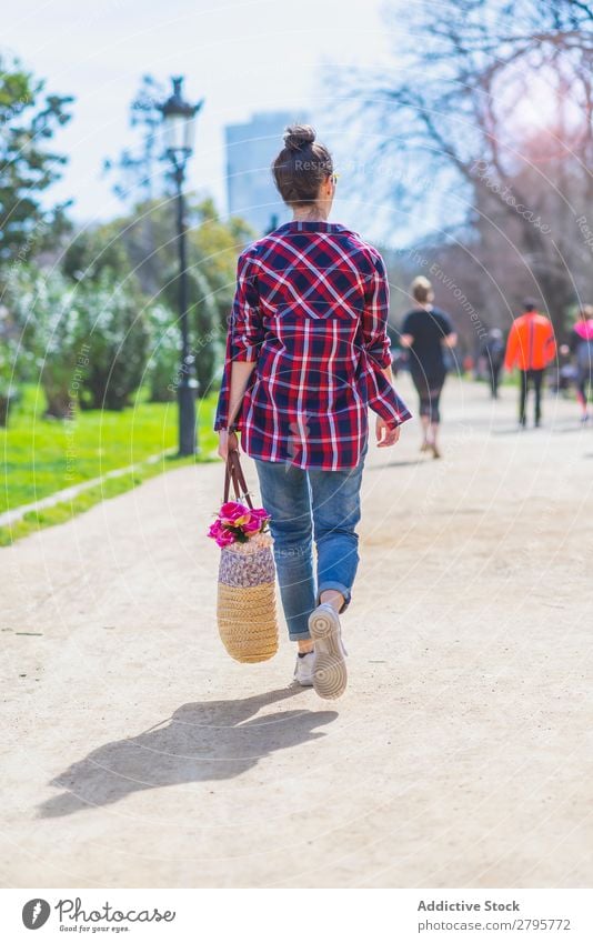 Rückansicht einer jungen Hipsterfrau, die an sonnigen Tagen in einem Park spazieren geht und dabei einen bösen Korb hält. Frau Junge Frau Schickimicki laufen