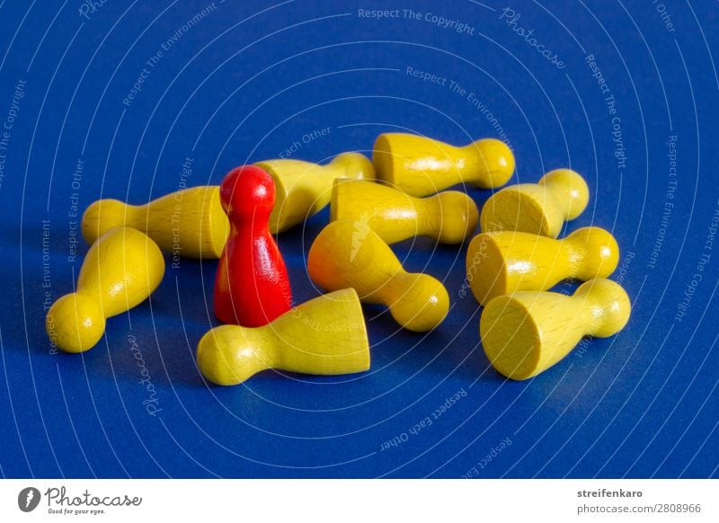 Eine rote Spielfigur steht inmitten von gelben liegenden Spielfiguren auf blauem Untergrund Menschengruppe Spielzeug Holz fallen kämpfen stehen Aggression