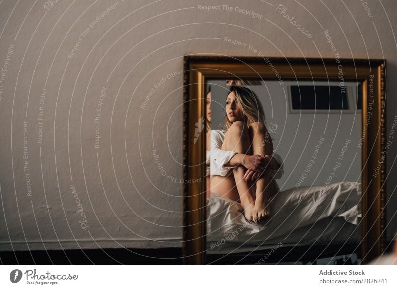 Das Spiegelbild einer Frau, die ihre Knie umarmt und auf einem Bett sitzt. Horizontale Innenaufnahme. umarmend Reflexion & Spiegelung Wegsehen Morgen heimwärts
