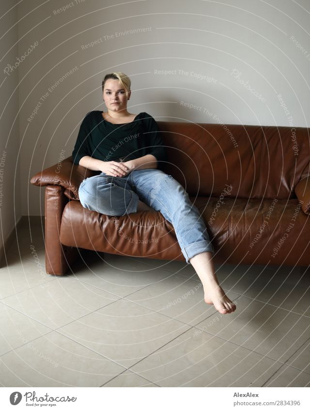 Junge Frau sitzt auf der Couch und streckt ein Bein heraus Stil Zufriedenheit Sofa Raum Jugendliche 18-30 Jahre Erwachsene Jeanshose Piercing Barfuß blond