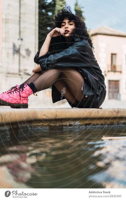 Junge Frau, die am Springbrunnen posiert. attraktiv Großstadt modisch lockig brünett Jacke Wasser Blick in die Kamera Mode Jugendliche schön hübsch Straße Model