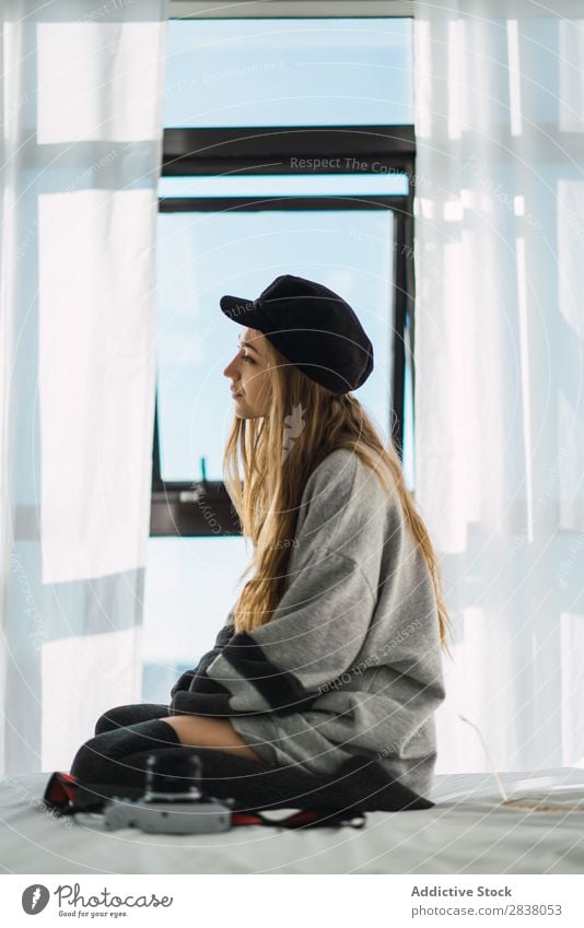 Stylisches junges Mädchen auf dem Bett sitzend Frau träumen Stil Erholung Pullover Hut genießen Körperhaltung Fenster weiß Fürsorge Schlafzimmer ruhen
