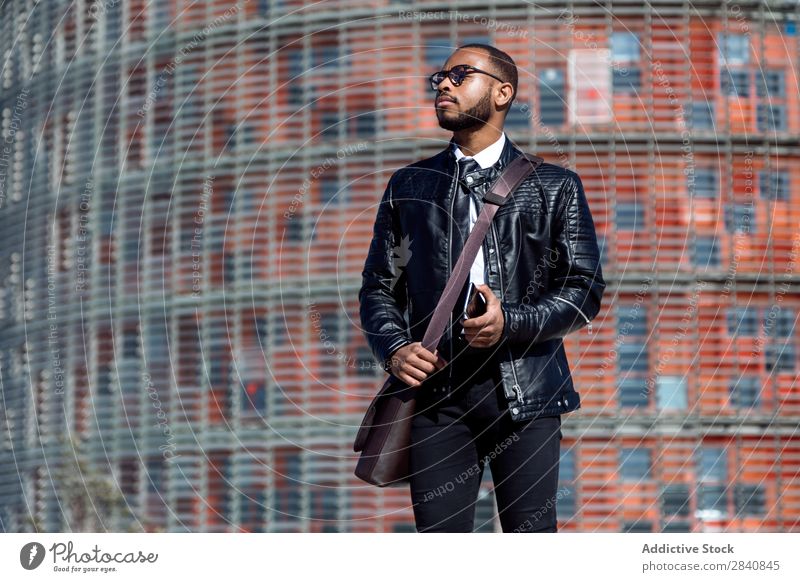 Modischer Mann in schwarzem Ledermantel und Brille, der sein Smartphone hält und auf den städtischen Hintergrund schaut. männlich gelungen Telefon stylisch
