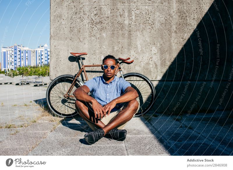 Lächelnder Mann mit Sonnenbrille am Fahrrad sitzend Fahrradfahren Pause schwarz Straße Beton Wand Sommer Jugendliche Lifestyle Großstadt Model Porträt