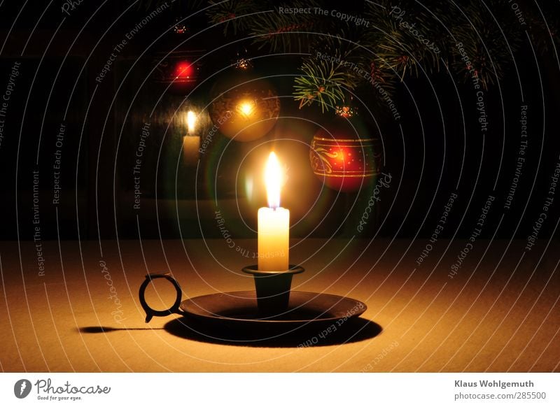 Eine einsame Kerze leuchtet, im Hintergrund Tannenzweige an denen Weihnachtsbaumkugeln im Kerzenschein glänzen. Die Kerzenflamme erzeugt Lensflares im Objektiv.