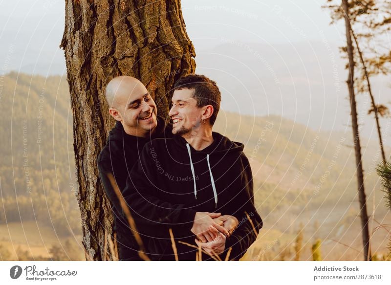 Lächelnde Männer, die sich am Baum im Wald auf einem Hügel umarmen. Homosexualität Paar Umarmen Liebe umarmend Tal malerisch Aussicht romantisch