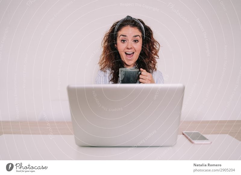 Junge Frau mit einem Becher in der Hand schaut überrascht auf ihren Laptop. Kaffee Lifestyle Glück Schreibtisch Tisch Arbeit & Erwerbstätigkeit Büro Business