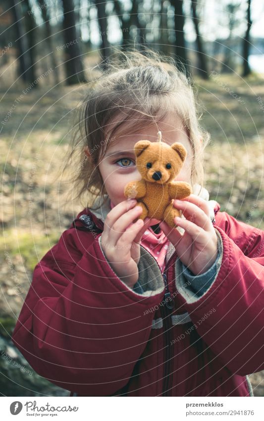 Kleines lächelndes glückliches Mädchen spielt mit ihrem kleinen Teddybär Spielzeug in einem Park an einem sonnigen Frühlingstag. Kind hält Teddy vor dem Gesicht und schaut in die Kamera mit roter Jacke
