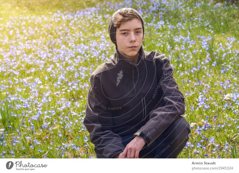 Porträt eines Jungen, der auf einer Frühlingswiese mit vielen Blumen kauert Mann männlich jung hockend Wiese im Freien Sonnenlicht blau Natur Gras grün Park