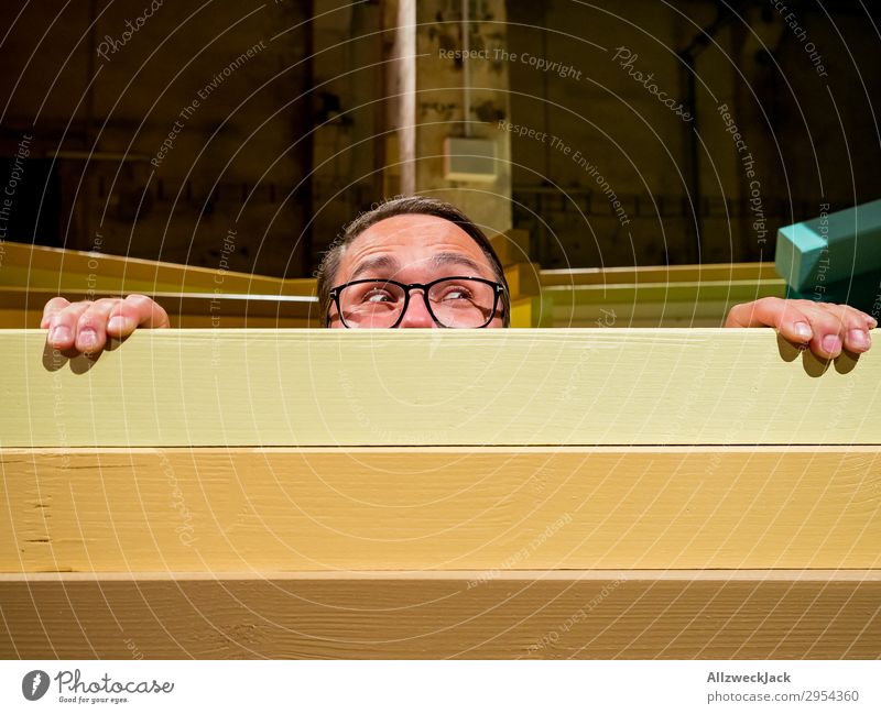 Junger Mann schaut neugierig über eine Absperrung Innenaufnahme 1 Mensch Kunstlicht Porträt Blick nach vorn Blick in die Kamera Brille Zaun Barriere Holzzaun