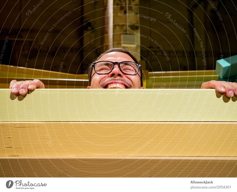 Junger Mann schaut über eine Absperrung Innenaufnahme 1 Mensch Kunstlicht Porträt Blick nach vorn Blick in die Kamera Brille Zaun Barriere Holzzaun spionieren