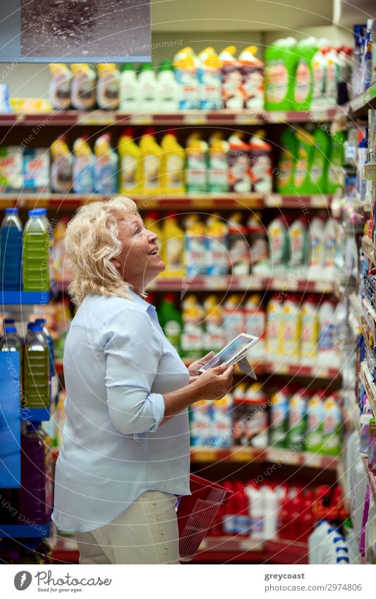 Eine lächelnde Frau mittleren Alters in einem hellblauen Hemd steht in einer Haushaltsabteilung eines Supermarktes. Sie hält ein Tablet und einen roten Einkaufskorb in ihren Händen. Eine Frau schaut sich die Regale an, auf der Suche nach etwas Bestimmtem