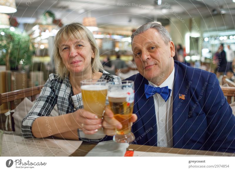 Ein Porträt eines Paares mittleren Alters, das dicht nebeneinander am Tisch sitzt. Der Mann trägt eine blaue Jacke und eine leuchtend blaue Fliege. Die Frau trägt ein kariertes Hemd. Beide erheben Gläser mit Bier und lächeln