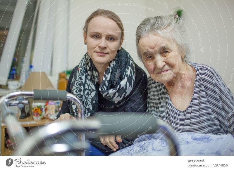 Ein Porträt einer jungen, blonden Frau, die dicht neben einer älteren Dame sitzt, deren Hand auf ihrem Arm liegt. Beide sind leicht lächelnd Mensch feminin