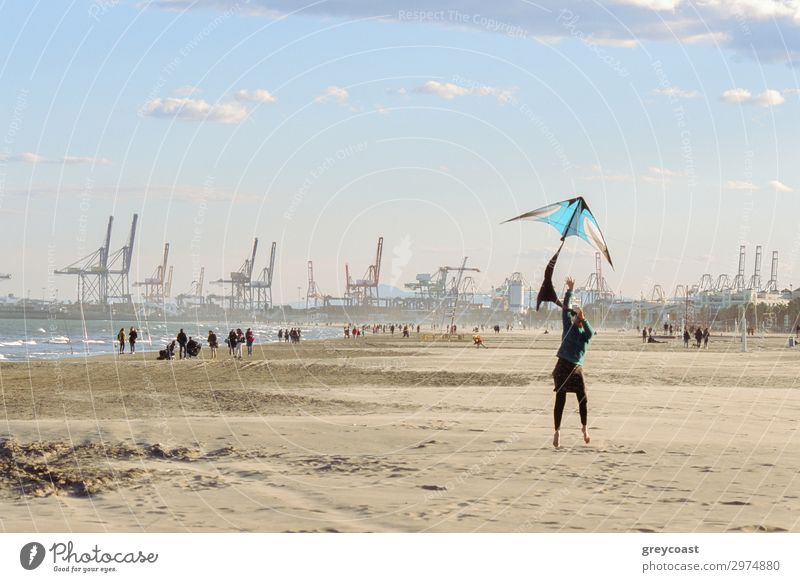 Eine Frau fliegt einen Drachen am Meeresufer. Promenierende Menschen und ein Hafen mit Kränen für Frachtschiffe im Hintergrund Freizeit & Hobby Strand Winter