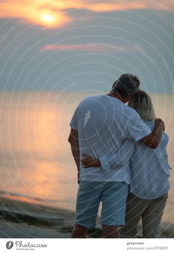 Ein Paar mittleren Alters, das sich umarmt und einen Sonnenuntergang am Meer beobachtet, wir sehen sie von hinten Ferien & Urlaub & Reisen Wellen Mensch