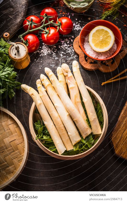 Spargel im Bambus-Dampfgarer auf dunklem, rustikalem Küchentisch mit Zutaten und Stäbchen, Draufsicht. Gesunde Ernährung und saisonales Kochen. Asiatisches Essen.