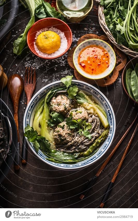 Asiatische Nudelschüssel mit grünem Gemüse, Bok Choy und Fleischbällchen auf dunklem Hintergrund mit Holzbesteck, Essstäbchen und scharfer Sauce, Ansicht von oben. Nahaufnahme. Gesundes asiatisches Essen. Konzept der asiatischen Küche