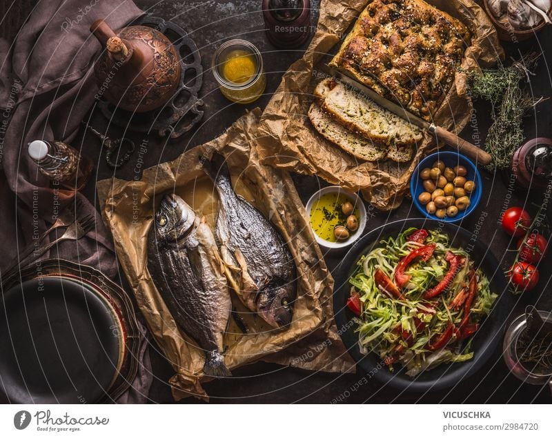 Mediterranes Mittag- oder Abendessen mit gebratenen Doradenfischen, hausgemachtem Focaccia-Brot, Olivenöl und Oliven, serviert auf einem rustikalen Tisch mit Geschirr und Küchenutensilien, Ansicht von oben.