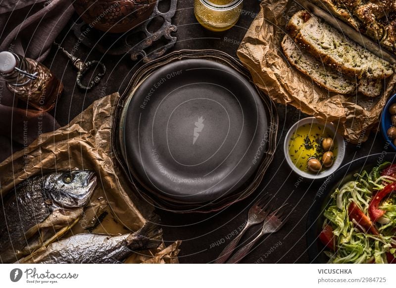 Essenshintergrund der mediterranen Küche mit Focaccia-Brot , gebratenen Doradenfischen, Salatschüssel und Olivenöl auf dunklem, rustikalem Küchentisch um den leeren Teller herum. Köstliches Konzept der mediterranen Küche.