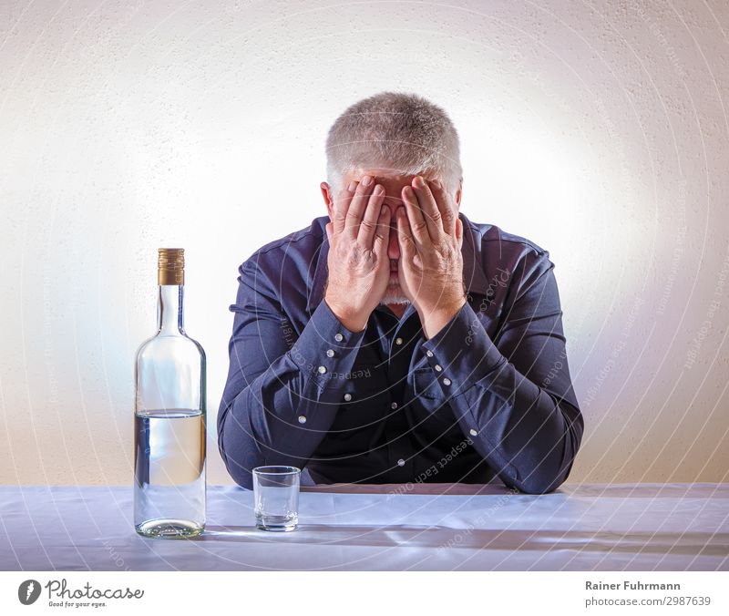 Ein Mann sitzt an einem Tisch und bedeckt mit den Händen sein Gesicht. Auf dem Tisch steht eine Flasche mit Alkohol und ein leeres Schnapsglas Wodka Alkoholiker