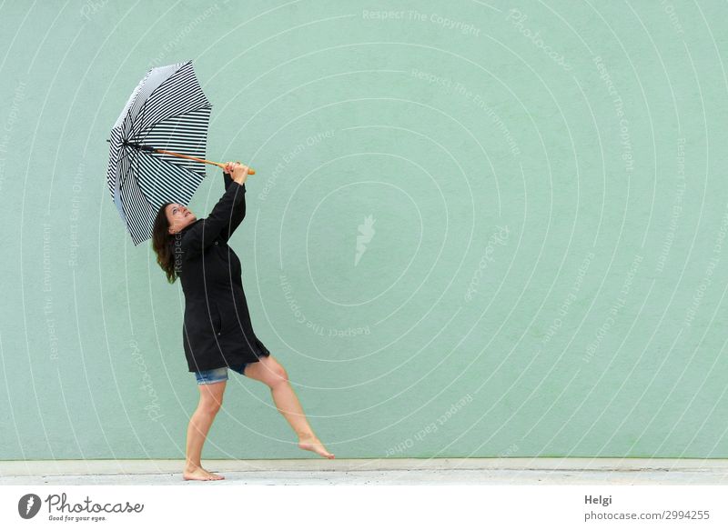 Frau mit schwarzem Mantel und kurzer Jeans tanzt barfuß mit einem Regenschirm in den Händen vor einer hellgrünen Wand Mensch feminin Erwachsene 1 45-60 Jahre