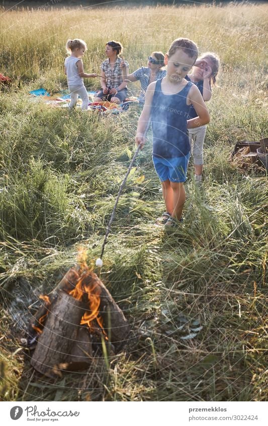 Kleiner Junge röstet Marshmallow über einem Lagerfeuer. Familie verbringt Zeit zusammen auf einer Wiese, nahe an der Natur. Eltern und Kinder sitzen auf einer Decke im Gras. Unverfälschte Menschen, echte Momente, authentische Situationen