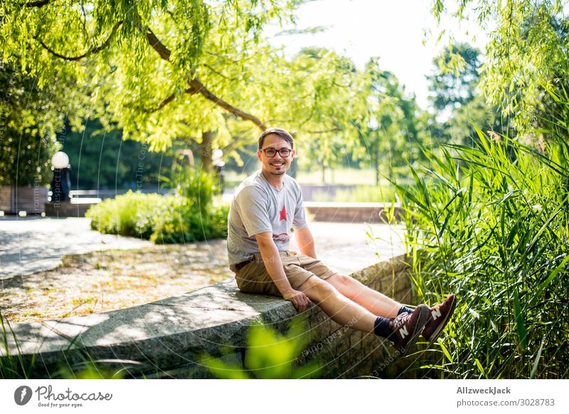 junger Mann sitzt auf einer Mauer und freut sich Schönes Wetter Wolkenloser Himmel Blauer Himmel grün Natur Baum Schilfrohr genießen Erholung Pause ausruhend