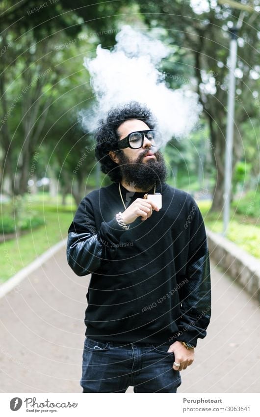 Junger Mann raucht eine elektronische Zigarette im Park. Lifestyle Sommer Jugendliche Erwachsene Sonnenbrille Vollbart rot Raps lässig Rauch Verdunstung vaping