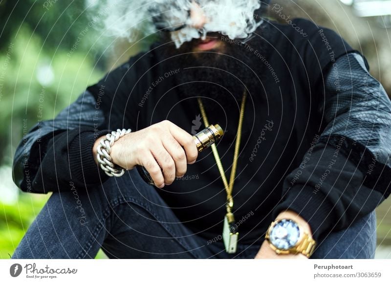 Mann mit Bart raucht eine elektronische Zigarette. Lifestyle Mensch Erwachsene Hand Natur Park Mode Bekleidung Sonnenbrille Hut Vollbart sitzen Coolness modern