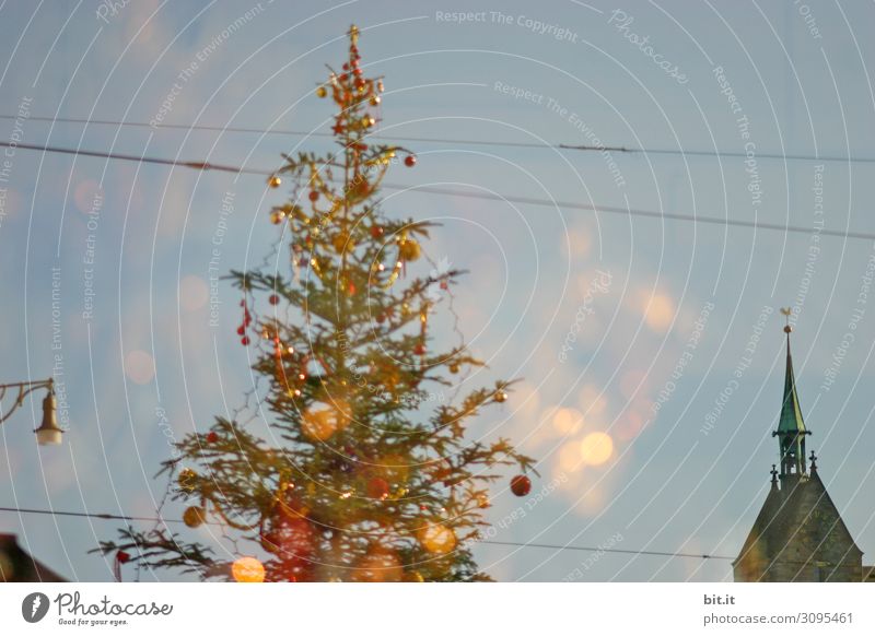 Kitschiger, skurriler, geschmückter Weihnachtsbaum steht zwischen Leitungen, Kirche, Lampe und Laterne in der Stadt und leuchtet. Lustige, abstrakte, leuchtende, witzige, urbane Weihnachtsstimmung mit Christbaum & Licht draussen vor blauem Himmel im Advent