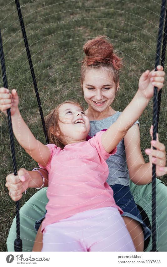 Teenager-Mädchen spielen mit ihrer jüngeren Schwester in einem Haus Spielplatz in einem Hinterhof. Glückliche lächelnde Schwestern haben Spaß auf einer Schaukel zusammen am Sommertag. Echte Menschen, authentische Situationen