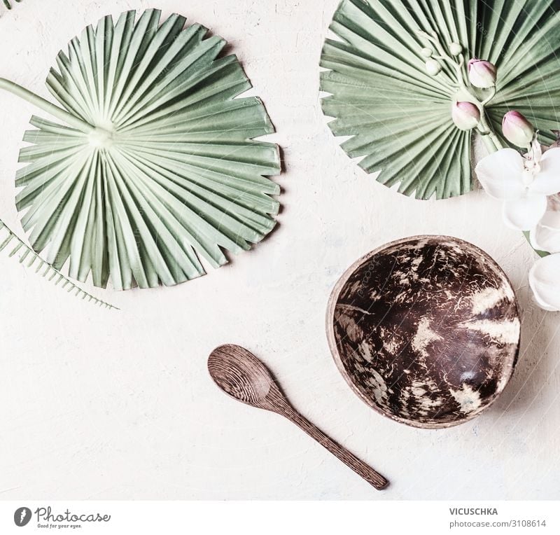 Leere Kokosnussschale mit Löffel auf weißem Schreibtischhintergrund mit tropischen Blättern, Ansicht von oben. Kopierraum für Ihr Design oder Produkt leer