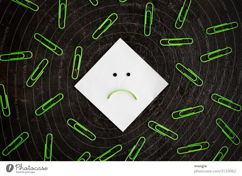 Ein Notizzettel als Smiley umgeben von vielen grünen Büroklammern Arbeit & Erwerbstätigkeit Büroarbeit Arbeitsplatz schwarz weiß Pessimist Mund Punkt negativ
