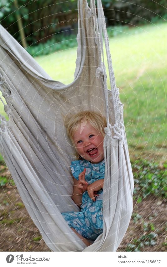 Kleines, süßes, blondes Mädchen sitzt, liegt glücklich und strahlend in einer Hängematte für Kinder im Garten und lacht herzhaft in die Kamera. Freizeit & Hobby