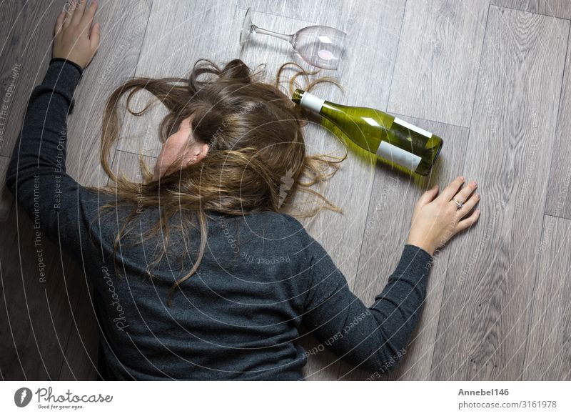 Betrunkenes alkoholisches junges Mädchen, das auf dem Boden liegt und sich krank fühlt, weil es Rotwein, ein leeres Glas und eine Flasche Knock-out getrunken hat