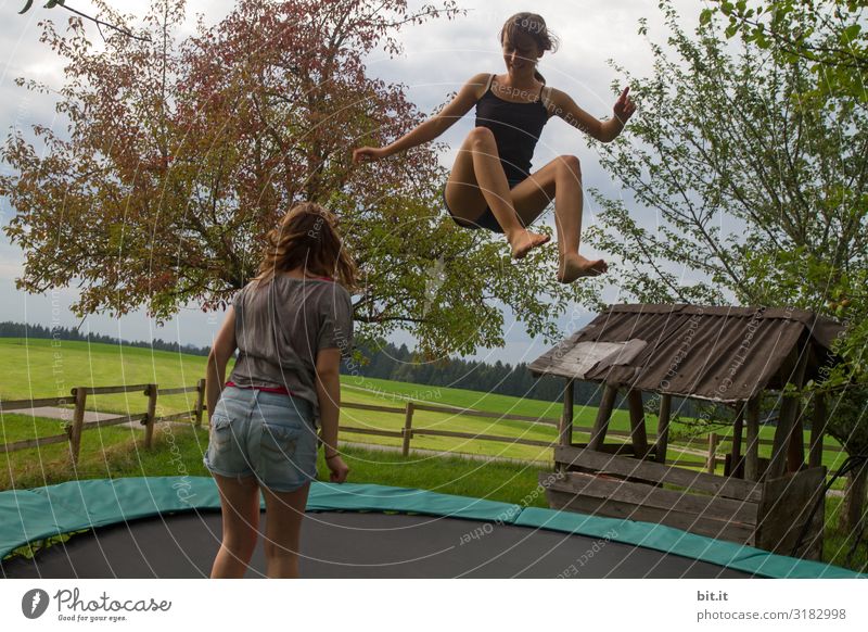 Zwei Mädchen hüpfen gemeinsam Trampolin, in der Natur. Eine fliegt hoch, die Schwester steht zum zuschauen, beobachten, lernen daneben. Sport Fitness Junge Frau