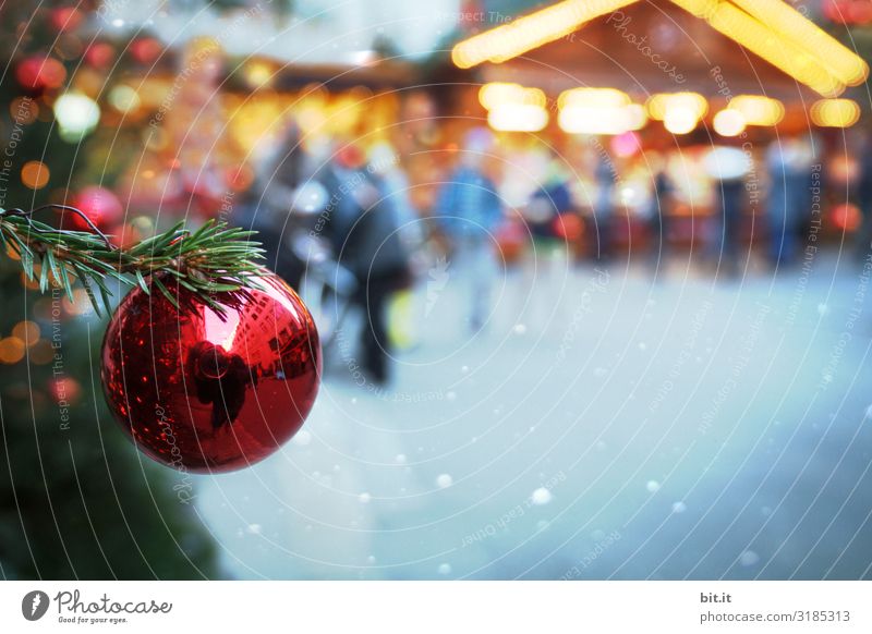 Unscharf l rote, glänzende Christbaumkugel, hängt an Tannenzweig in grün, auf beleuchteten Weihnachtsmarkt. Weihnachtsstimmung mit Christbaum auf Weihnachtsmarkt, beleuchtet mit viel Licht, in der Dämmerung. Abendstimmung auf Christkindlesmarkt mit Schmuck