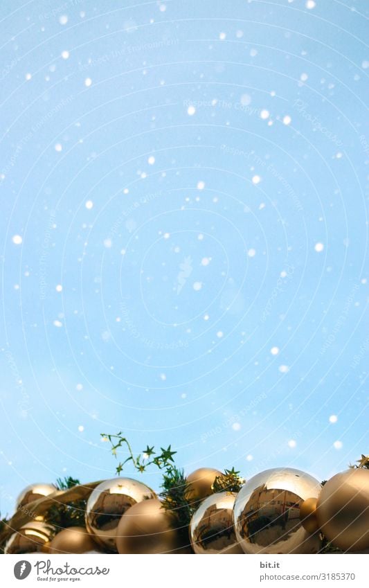 Glänzende, goldene Weihnachtskugeln im Schneefall vor blauem Himmel. Viele, funkelnde Christbaumkugeln im Schnee. Schneeflocken im Advent mit Weihnachtsdekoration, Verzierung unten. Kitschige Kugeln zur winterlichen, adventlichen, kalten Weihnachtszeit.