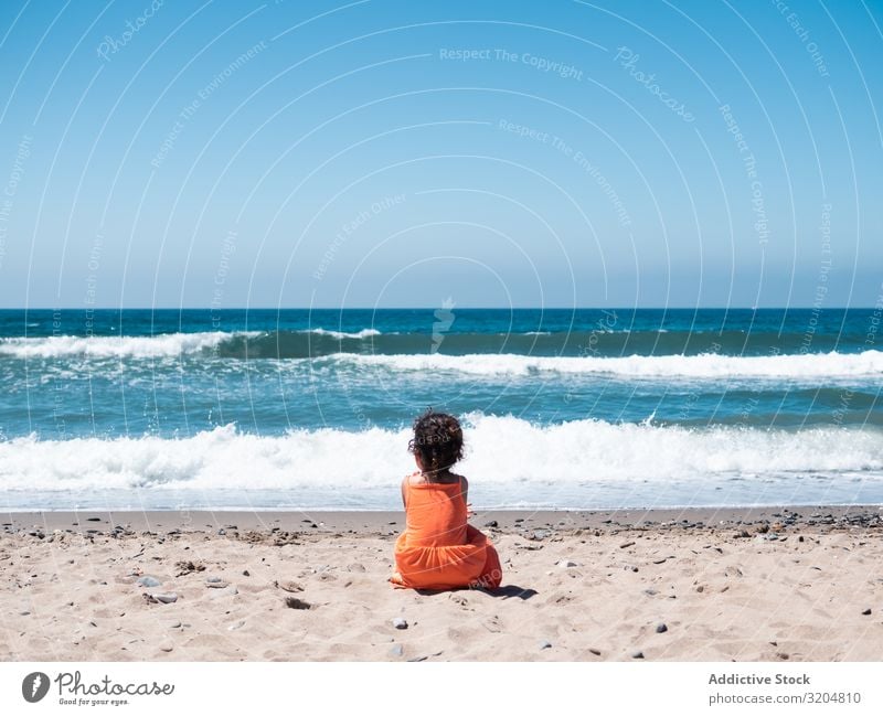 Kleines Mädchen sitzt am Strand und bewundert die Aussicht Kind bewundernd sitzen Seeküste Sand Meer Himmel klein Frau Orange Kleid Blick schön Sommer winken