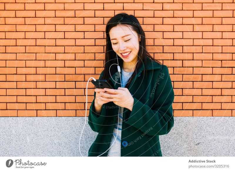 Asiatische Frau hört Musik in der Nähe einer Ziegelmauer hören PDA benutzend Wand anlehnen Backstein Straße Großstadt asiatisch urwüchsig Lifestyle