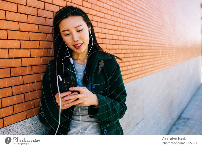 Asiatische Frau hört Musik in der Nähe einer Ziegelmauer hören PDA benutzend Wand anlehnen Backstein Straße Großstadt asiatisch urwüchsig Lifestyle