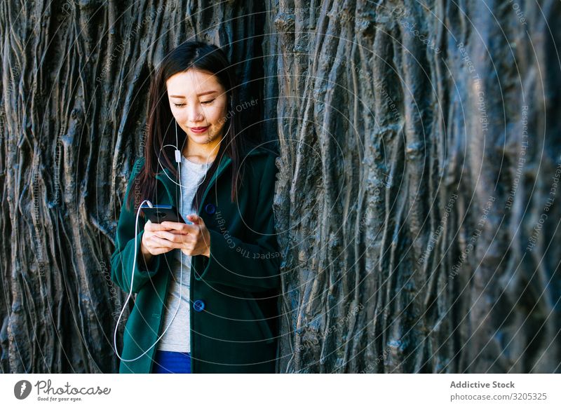 Asiatische Frau hört Musik in der Nähe einer rauen Wand hören PDA benutzend anlehnen Wurzel Baum Straße Großstadt asiatisch urwüchsig Relief Oberfläche