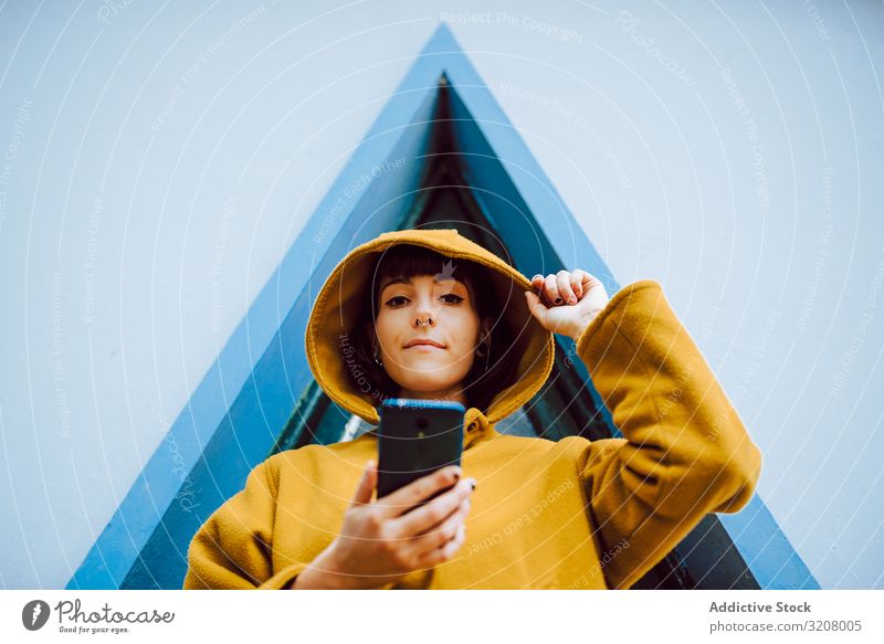 Frau mit Smartphone gegen Dreiecksfenster benutzend ausrichten Kapuze Fenster Gebäude Wand grau jung lässig Jacke warm Außenseite Mantel geometrisch Form Dame