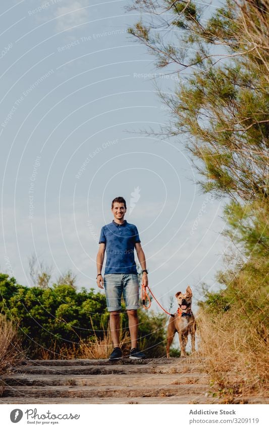 Glücklicher erwachsener Mann, der mit seinem Hund auf dem Land spazieren geht Spaziergang lässig Weg heiter anleinen braun ländlich Landschaft hölzern Baum Grün