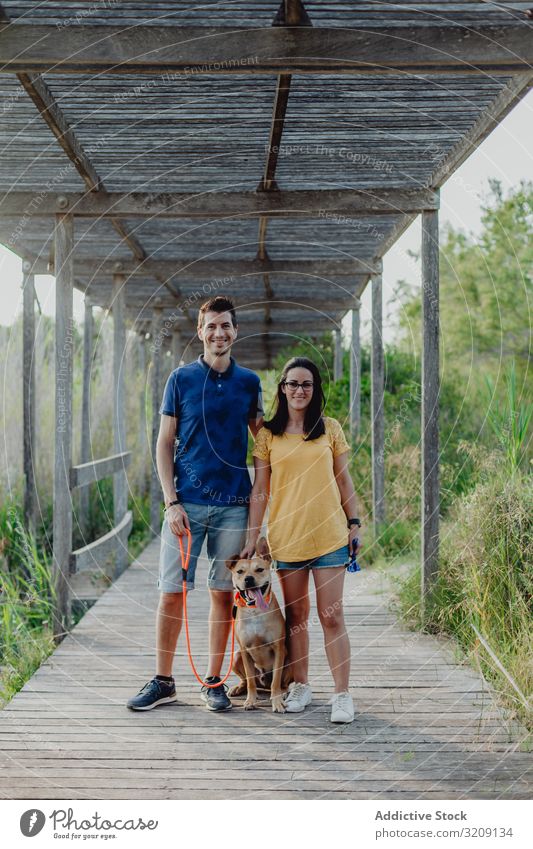 Glückliches erwachsenes Paar mit Hund auf dem Land stehend spielen Landschaft lässig heiter anleinen braun ländlich hölzern Frau Mann Baum Stehen Terrasse Grün