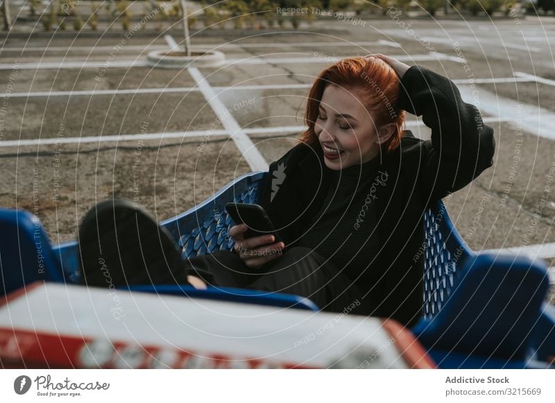 Lächelnde Frau mit Smartphone im Einkaufswagen auf dem Parkplatz Selfie attraktiv Selfie nehmen jung schön lässig heiter klug Netzwerk Telefon modern Stehen