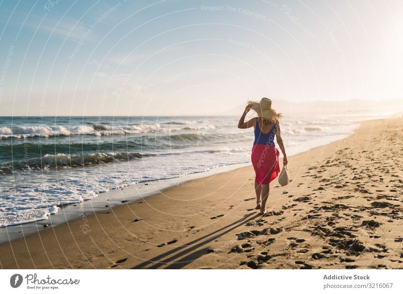 Frau hält Hut und geht am Meer entlang Strand sandig Badebekleidung pareo Wasser laufen Sommer genießend Freizeit sportlich Körper Glück schlank Seeküste wellig