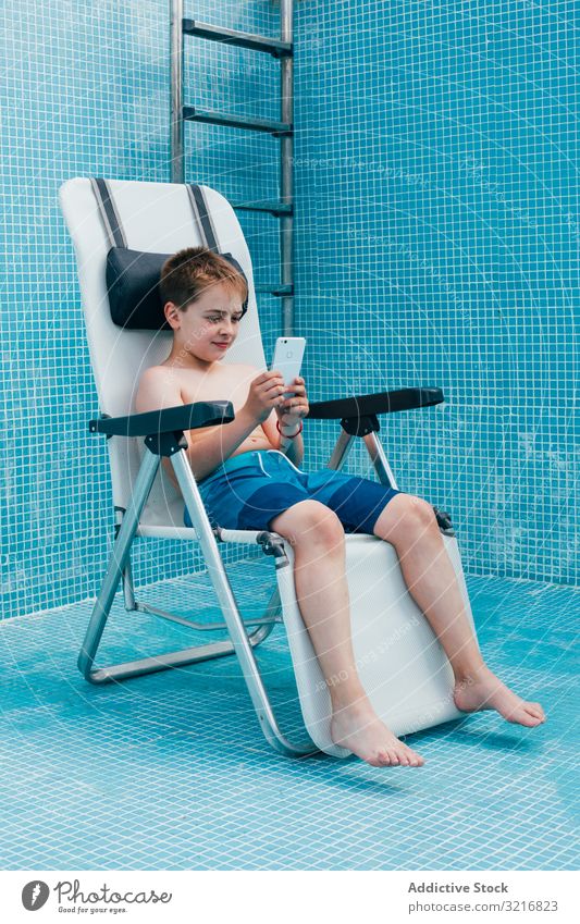 Junge mit Smartphone auf dem Boden eines leeren Schwimmbeckens sitzend Pool Sitzen Gesäß Kind aussruhen Stuhl Fliesen u. Kacheln dekoriert Technik & Technologie